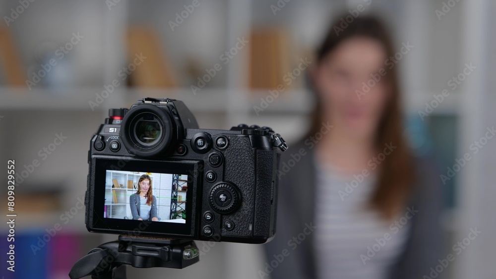 Pretty female blogger recording video with camera at home, focus is on camera screen on tripod. Woman speaking in front of camera for her vlog