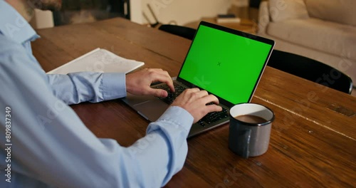 A man in formal clothes uses a laptop with green screen sitting at a table indoors. An unrecognizable person