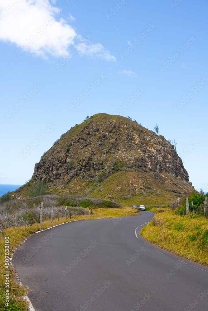 Road curves around mountain rocks on the ocean with blue sky background, unrecognized cars parking in front.