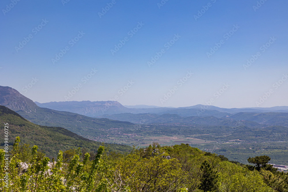 vista dettagliata, dall'alto, di giorno, in estate, sotto un cielo sereno e color azzurro, del paesaggio naturale tra pianura, collina e montagna nell'area ovest della Slovenia