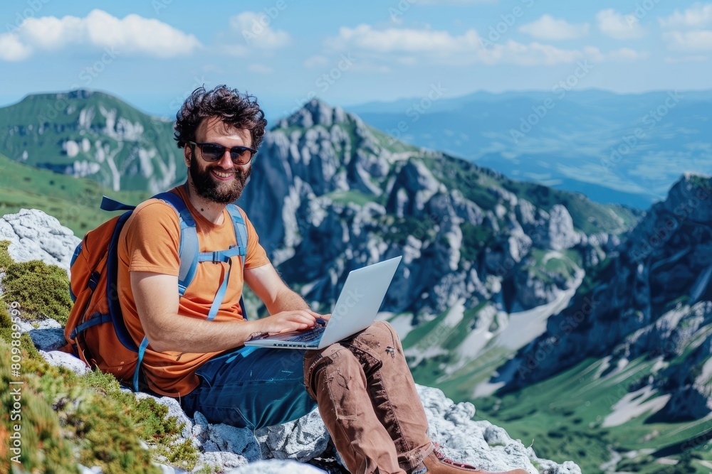 A digital nomad smiling while working on his laptop, seated on a mountain peak with a breathtaking view of the landscape around.