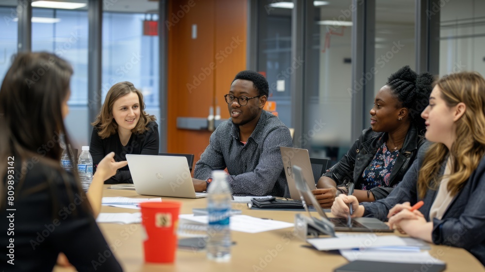 A diverse group of individuals is seated around a table, each engaged with a laptop, working or collaborating on a project. A diverse group of individuals is seated around a table, each engaged with a laptop, working or collaborating on a project.