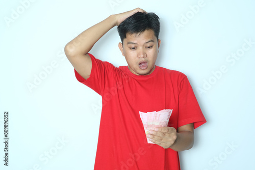 Young Asian man showing shocking expression while looking to paper money. Wearing red t-shirt photo