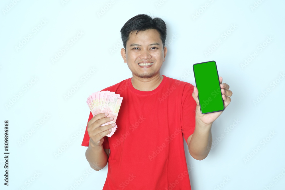 Young Asian man smiling happy while showing green screen mobile phone and holding paper money. Wearing red t-shirt Young Asian man smiling happy while showing green screen mobile phone and holding paper money. Wearing red t-shirt