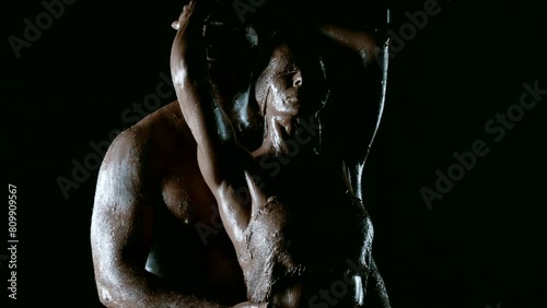 In the darkness on a black background, a stunning scene of passion and devotion unfolds, where a man with a muscular physique and his beautiful wife, decorated with clay