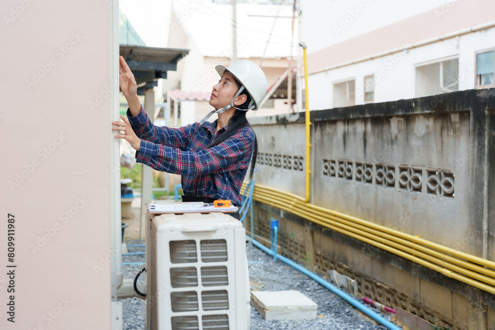 Asian woman in plaid shirt, white helmet, examines outdoor air conditioning unit with focus, against residential backdrop. inspects house. happy at work, Inspectors inspect the completed house.