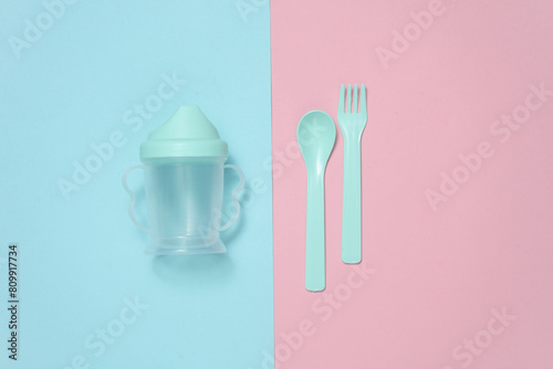 Drinking bowl and plastic fork and spoon for baby on a blue and pink background. Top view