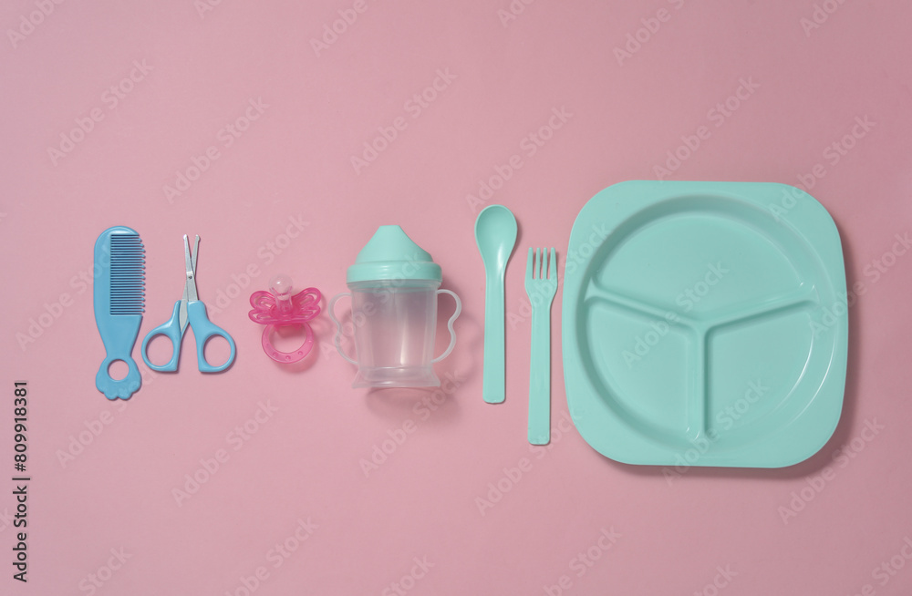 Accessories for caring and feeding a baby on a pink background. Scissors, comb, drinking bowl, plate and fork with spoon on pink background. Top view