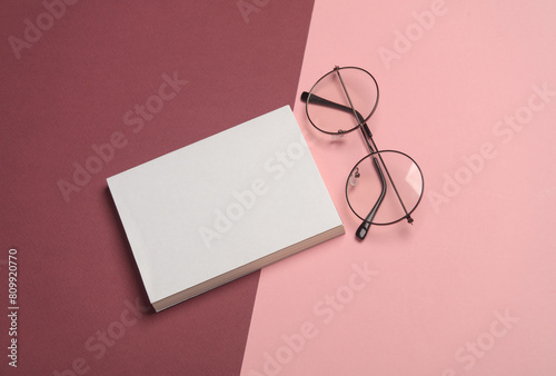 Stylish eyeglasses and book or notebook with white blank cover on burgundy pink background.