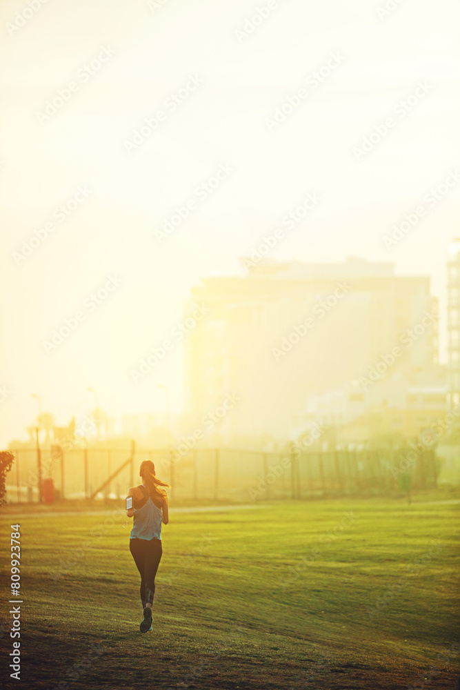 Fototapeta premium Woman, running and back on grass for exercise with mock up space in sky with buildings in city. Girl, person and athlete with training, workout and speed in nature on field for wellness in morning