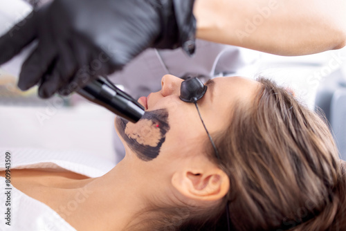 Beautician holding beauty laser near woman face covered with carbon mask