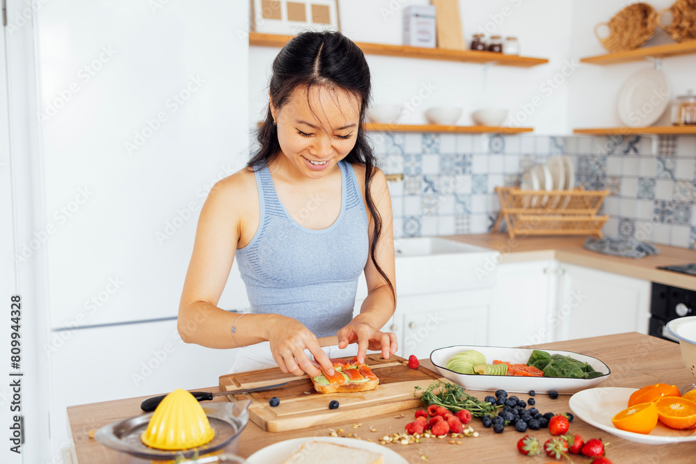 Young attractive Asian woman prepares delicious sandwich with fish and avocado. Smiling lovely Korean girl in casual clothes prepares breakfast in kitchen at home. Young attractive Asian woman prepares delicious sandwich with fish and avocado. Smiling lovely Korean girl in casual clothes prepares breakfast in kitchen at home.