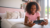 Teenage Girl Lying On Bed At Home Playing With Handheld Gaming Device