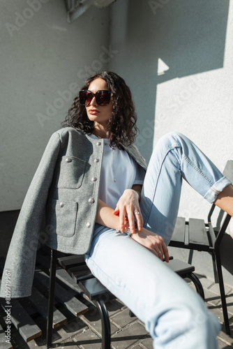 Cool beautiful fashionable stylish woman with curly hair with eyewear in fashion clothes with jeans, white T-shirt and blazer sitting and posing on the street