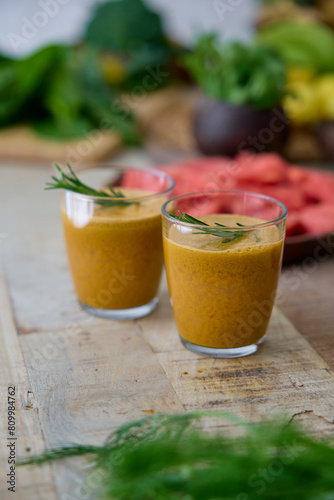 This image showcases two glasses of freshly blended green smoothie, garnished with rosemary, surrounded by an assortment of fresh vegetables and fruits like spinach and watermelon.