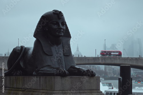 An Egyptian sphinx on the banks of the River Thames and a red bus on the bridge on a foggy day. Waterloo Bridge, London, United Kingdom