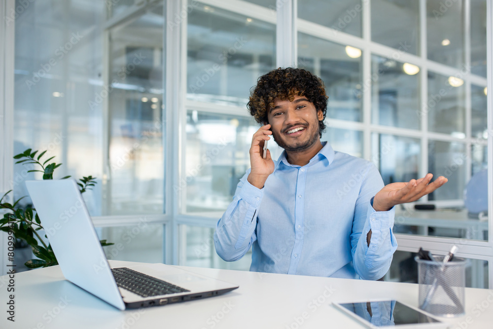 Obraz premium Cheerful young man with curly hair, wearing a light blue shirt, communicates on a smartphone while working at a white desk in a bright office environment.