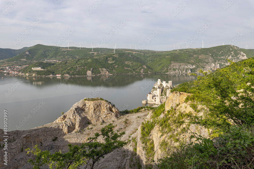 serene lakeside scene dominated by a medieval castle perched atop a cliff. The surrounding landscape is dotted with wind turbines serene lakeside scene dominated by a medieval castle perched atop a cliff. The surrounding landscape is dotted with wind turbines