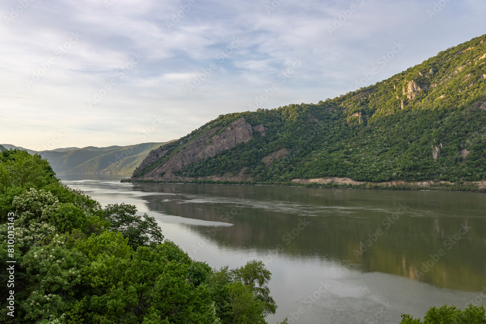 A tranquil river scene with a forested mountain on one side, reflecting in the calm water, under a cloudy sky