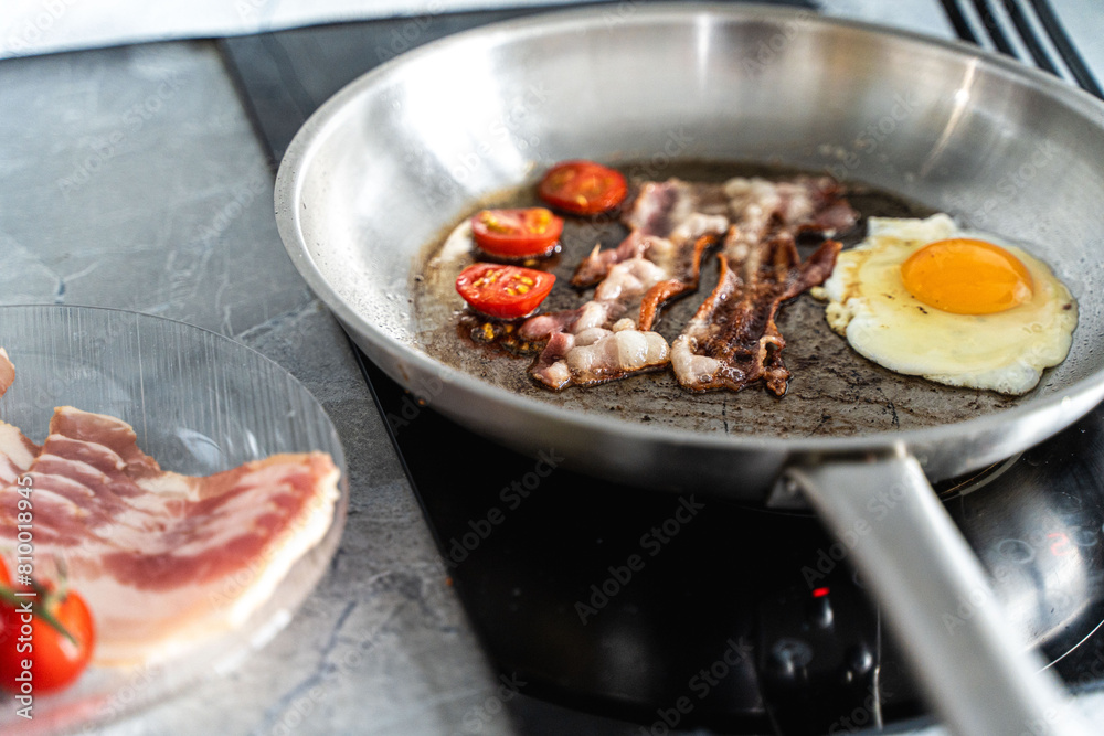 A pan of scrambled eggs, bacon and cherry tomatoes sits on the stove in the kitchen next to a plate of fresh tomatoes and raw bacon. A pan of scrambled eggs, bacon and cherry tomatoes sits on the stove in the kitchen next to a plate of fresh tomatoes and raw bacon.