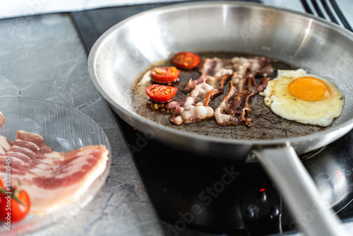 A pan of scrambled eggs, bacon and cherry tomatoes sits on the stove in the kitchen next to a plate of fresh tomatoes and raw bacon.