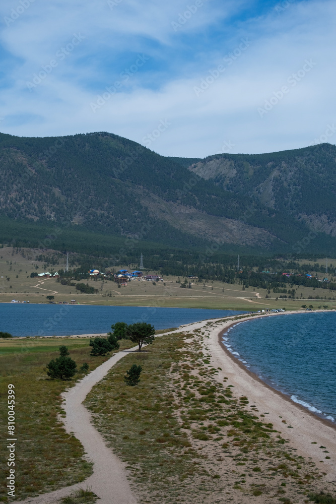 Cape Uyuga on Lake Baikal. Spit, part of land, protrudes deep into blue water surface on sides. Side top view. Waves hitting rocky shore, trees growing on the shore, forested mountains in background