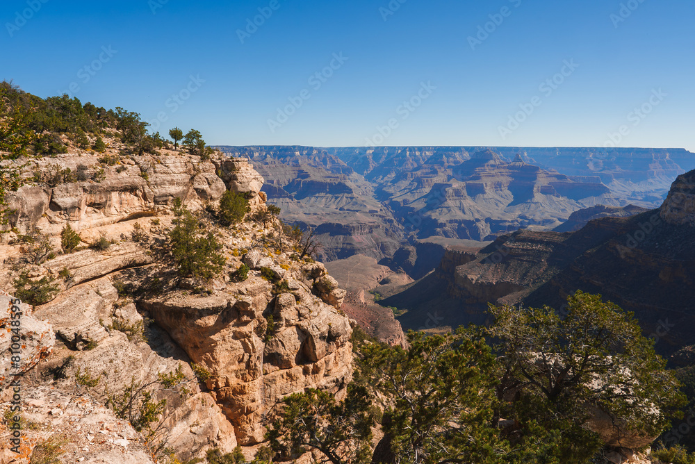 Scenic view of the Grand Canyon under a clear blue sky in Arizona, USA. Rugged, rocky terrain with intricate red, brown, and beige rock formations fading into the horizon.