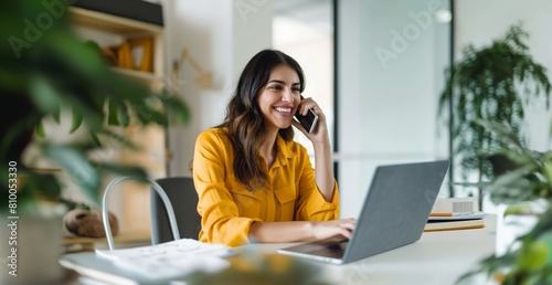 A woman is talking on her cell phone while sitting at a desk with a laptop. She is smiling and she is enjoying her conversation,wearing yellow clothes