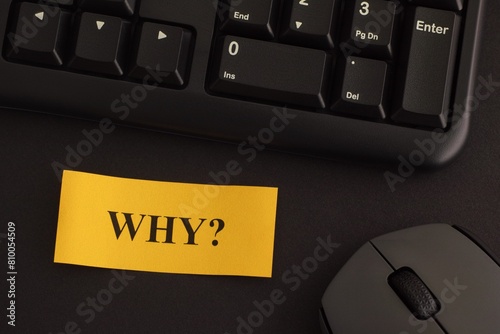 A paper note with the question Why? on it on a desk with a black keyboard and a grey wireless mouse. Close-up.