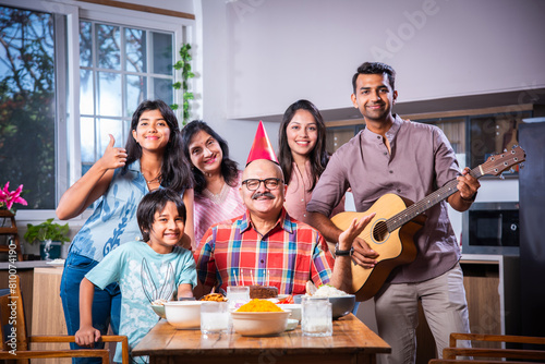 Indian asian grandparents with Family celebrating birthday by blowing candles on cake at home on dining table.