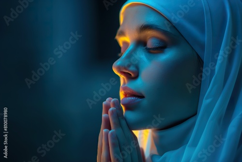 A young nun in a blue veil is devoutly praying against a dark blue background. photo