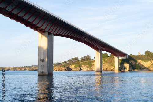 A bridge spans a body of water, with a view of the shoreline in the background. The bridge is red and white, and the water is calm