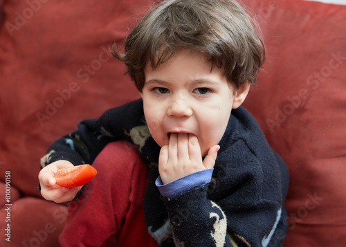 cute young boy in a sweater is eating vegetables on a red couch in the livingroom photo