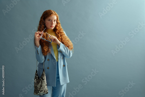 Fashionable redhead woman wearing elegant blue suit blazer, trousers, holding silver metallic purse. Studio fashion portrait. Copy, empty, blank space for text