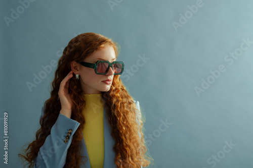 Fashionable redhead confident woman wearing trendy green sunglasses, silver earrings, yellow blouse, blazer, posing on blue background. Studio fashion portrait. Copy, empty, blank space for text