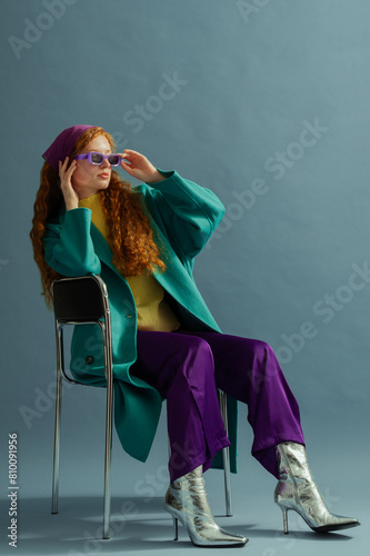 Fashionable redhead woman wearing purple headscarf, sunglasses, wide leg trousers, yellow blouse, green midi coat, silver ankle boots, posing on blue background. Studio full-length fashion portrait