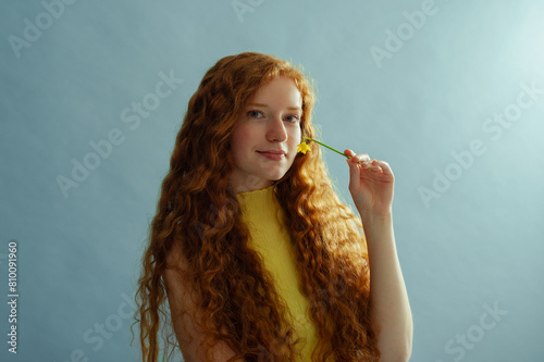 Beautiful happy smiling redhead freckled woman with long curly hair, holding daffodil, posing on blue background. Close up studio portrait. Copy, empty, blank space for text