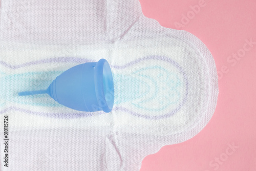 Blue menstrual cup is placed on sanitary pad, depicted against contrasting pink background to symbolize feminine hygiene and eco-friendly alternatives.