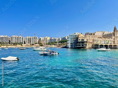 view of a bay on the island of Malta with some boats docked on the Mediterranean Sea and city buildings in the background on a sunny summer day 