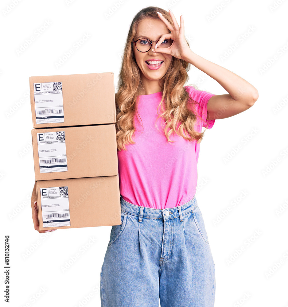 Young caucasian woman with blond hair holding delivery package smiling happy doing ok sign with hand on eye looking through fingers