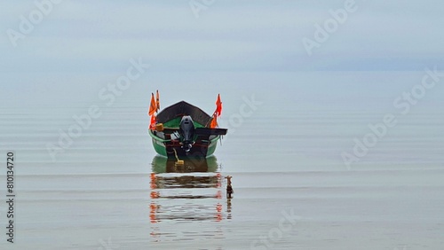 Boat with red flags anchored on calm sea surface against a gray cloudy sky