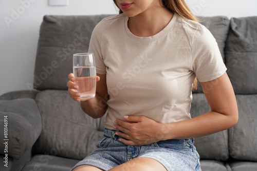 Costiveness. Unreconizable young woman suffering from abdominal pain holds a glass of water in her hand at home.