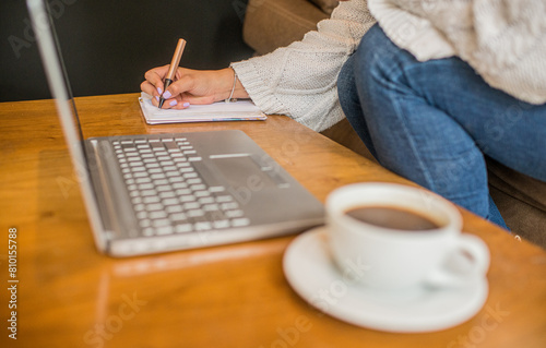 primer plano de manos de mujer tomando apuntes en su libreta al lado del computador portátil y un café 
