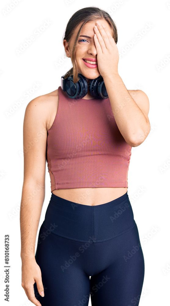 Beautiful caucasian young woman wearing gym clothes and using headphones covering one eye with hand, confident smile on face and surprise emotion. Beautiful caucasian young woman wearing gym clothes and using headphones covering one eye with hand, confident smile on face and surprise emotion.