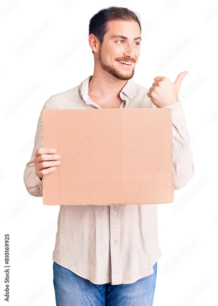 Young handsome caucasian man holding empty cardboard banner pointing thumb up to the side smiling happy with open mouth Young handsome caucasian man holding empty cardboard banner pointing thumb up to the side smiling happy with open mouth