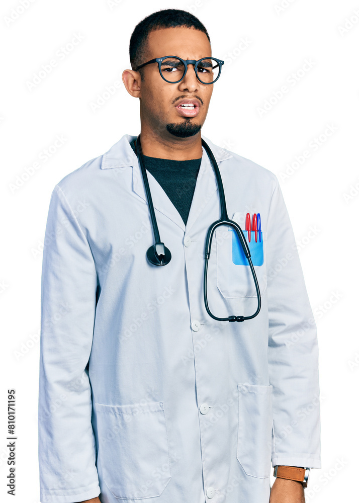 Fototapeta premium Young african american man wearing doctor uniform and stethoscope in shock face, looking skeptical and sarcastic, surprised with open mouth