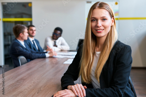 Confident blonde businesswoman leads meeting at modern office. Young caucasian female boss sits at table with multiethnic team. Colleagues discuss strategy, collaboration in corporate environment. photo