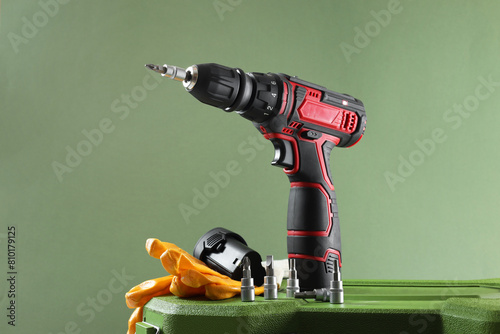 Electric screwdriver, drill bits, battery and gloves on case against pale green background photo