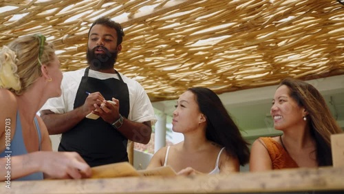 Happy diverse female friends choose, order food and drinks at cafe terrace on Bali. Multiracial women read menu in a restaurant. Indian smiling waiter takes orders from clients and recommends menu.