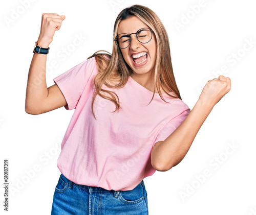 Hispanic young woman wearing casual t shirt and glasses very happy and excited doing winner gesture with arms raised, smiling and screaming for success. celebration concept. photo